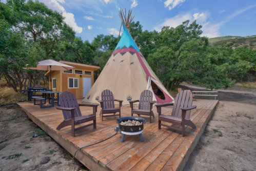 A wooden deck with a fire pit and chairs, featuring a colorful teepee and surrounded by trees under a blue sky.